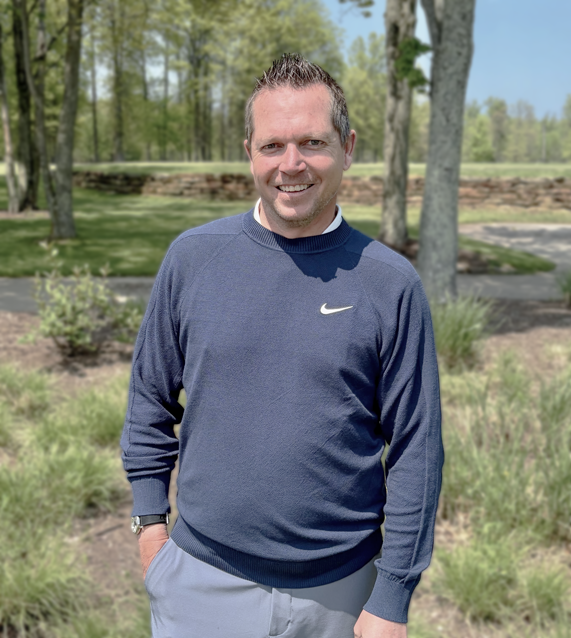 Smiling man in a navy Nike sweater standing in a park with trees in the background on a sunny day.