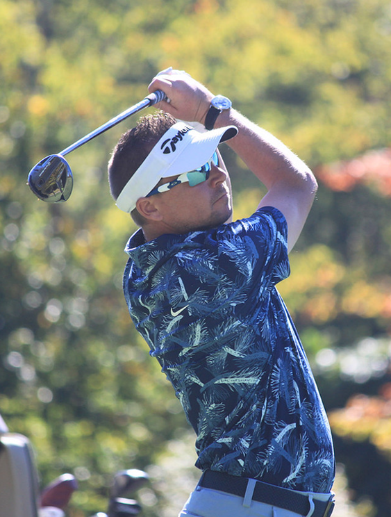 Golfer mid-swing on a sunny golf course, wearing a white visor and blue tropical-pattern shirt with sunglasses.