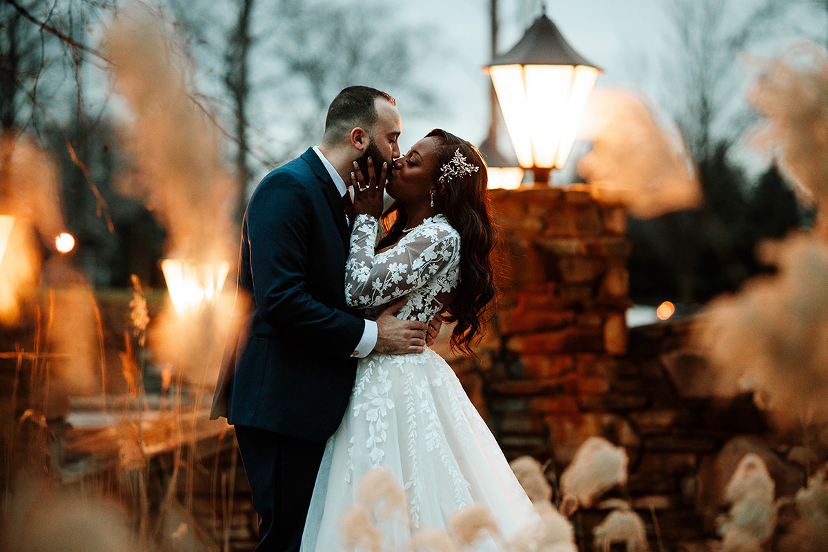 Couple in wedding attire share a kiss outdoors at dusk beside a stone wall and glowing lanterns.