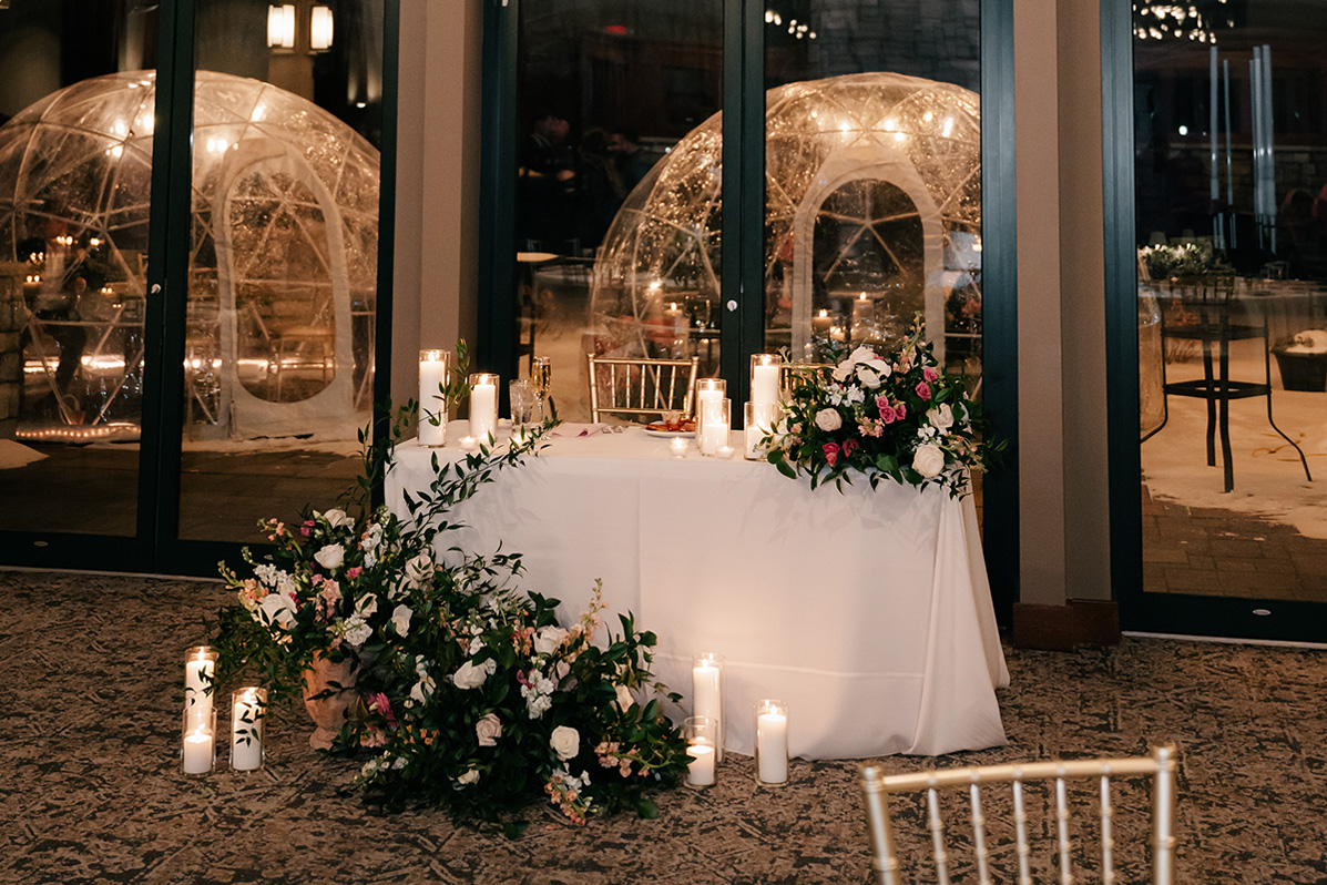 Head table with white cloth, tall candles, and lush floral arrangements in a dim banquet hall setting.