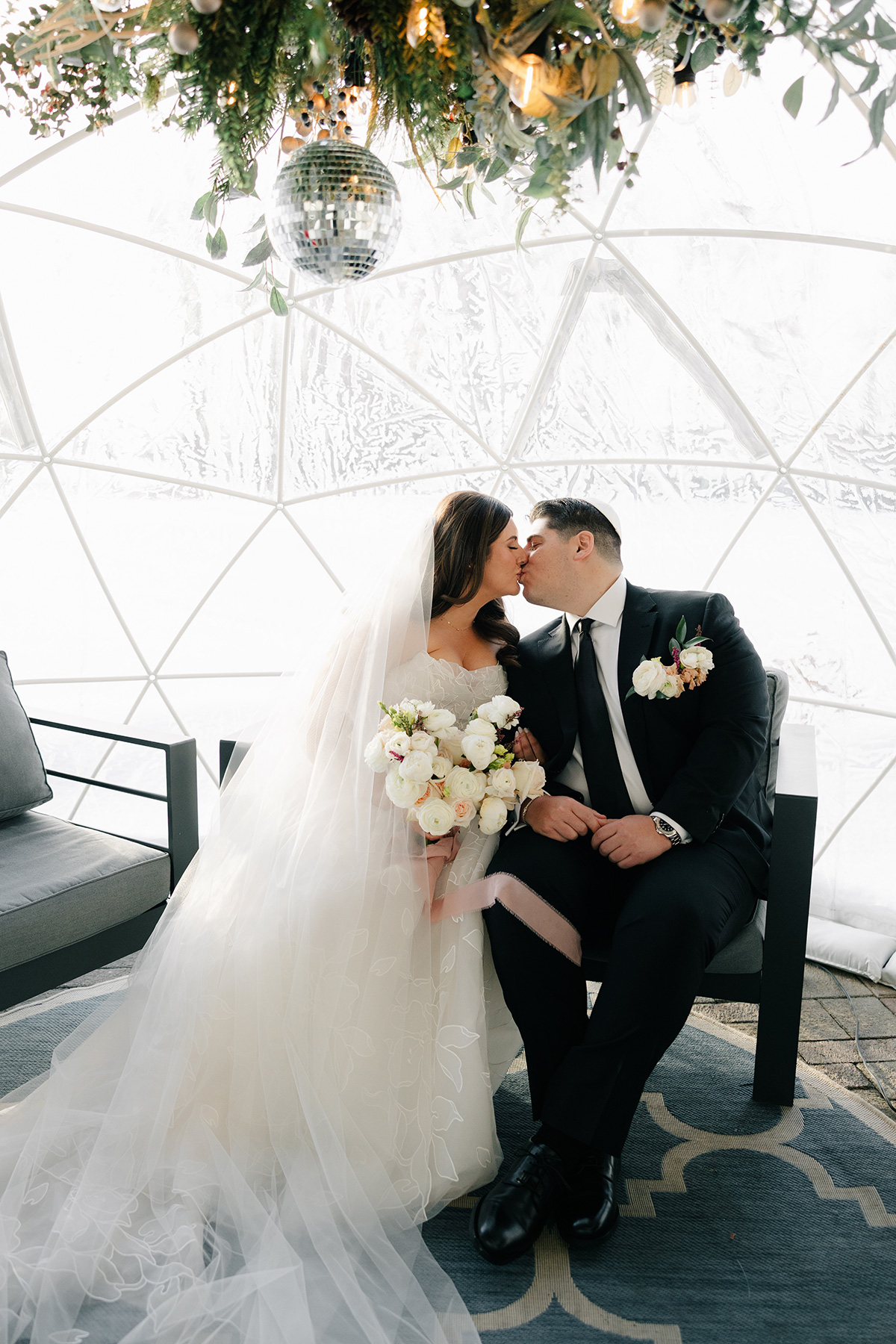 Bride in a white gown and veil kisses a groom in a black suit inside a glass geodesic dome, holding a white bouquet of roses amid greenery and a hanging disco ball
