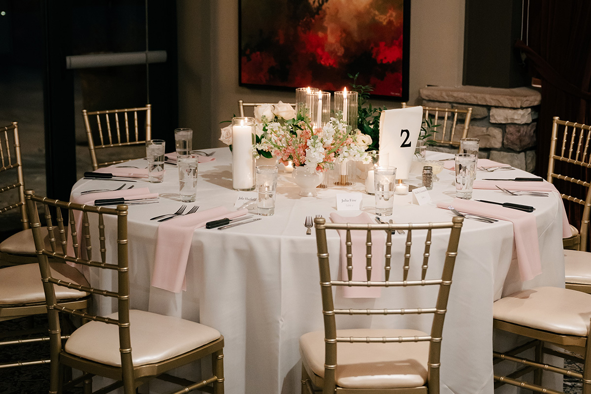 Round banquet table set for a formal event with pink napkins, gold chairs, a floral centerpiece, and tall candles, table number 2 visible