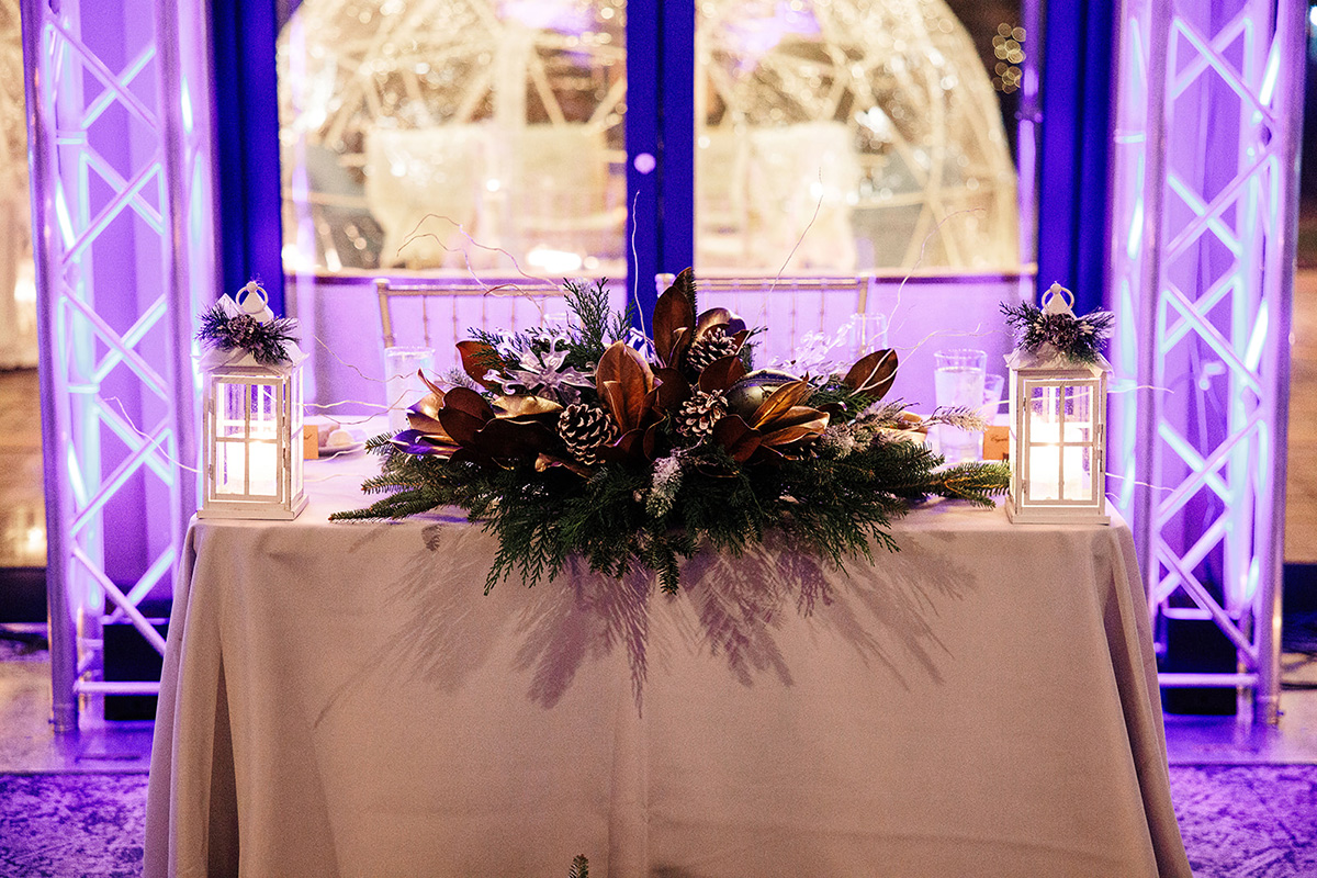 Table centerpiece of evergreen branches, pinecones, and bronze leaves with two white lanterns and candles; purple lighting behind.