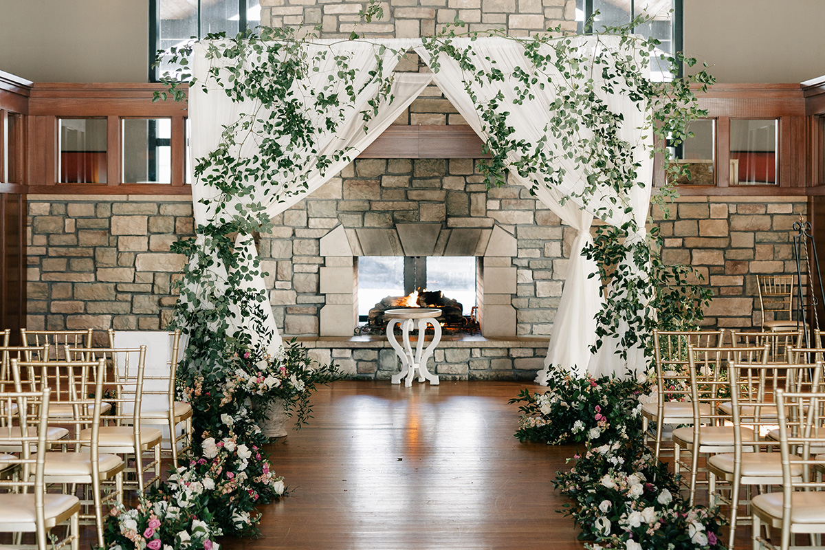 Stone-fireplace ceremony arch with white drapes and greenery, floral aisle, and gold chairs for a wedding venue.