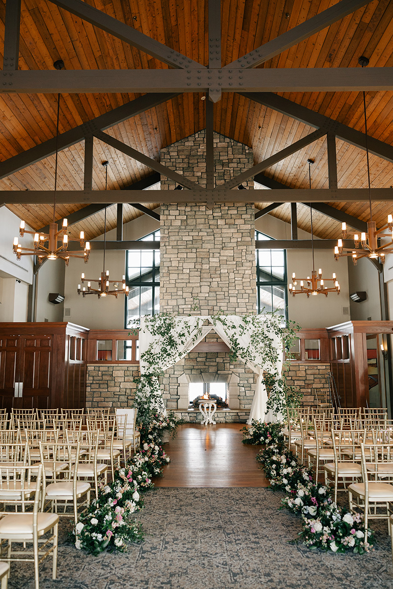 Indoor wedding ceremony space with a stone fireplace altar framed by a floral arch and rows of gold chairs along a flower-adorned aisle.