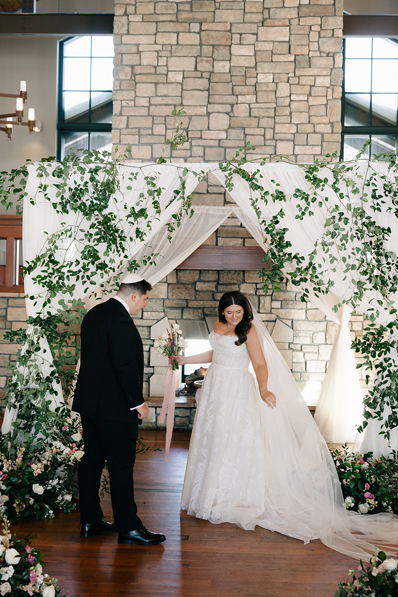 Bride in a white wedding gown and veil stands under a leafy floral arch, holding a bouquet, as the groom in a black suit looks on in a stone chapel setting.