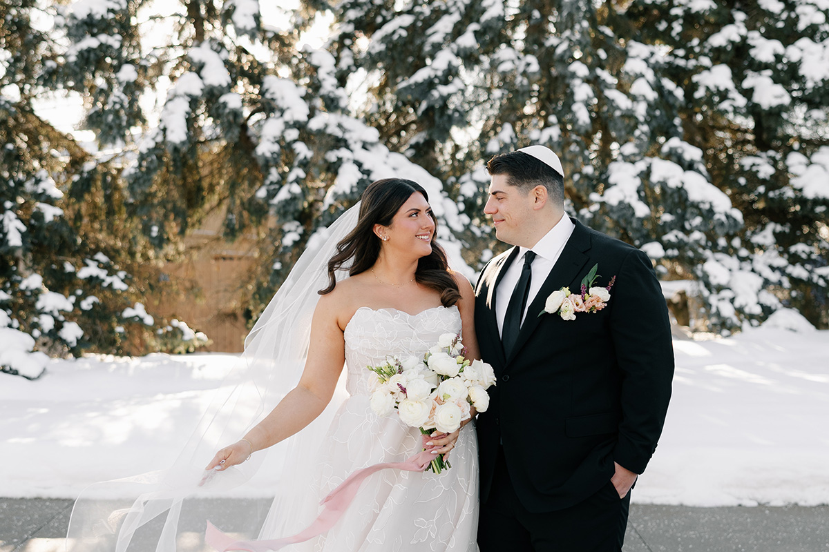 Bride in a white wedding dress with veil holds a white bouquet as a groom in a black suit smiles at her in a snowy outdoor setting.