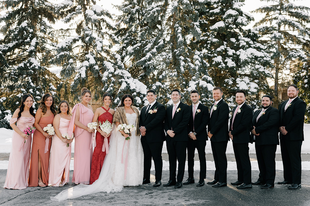 Bride and groom with a line of bridesmaids in pink dresses and groomsmen in black suits outdoors in a snowy setting, holding bouquets.