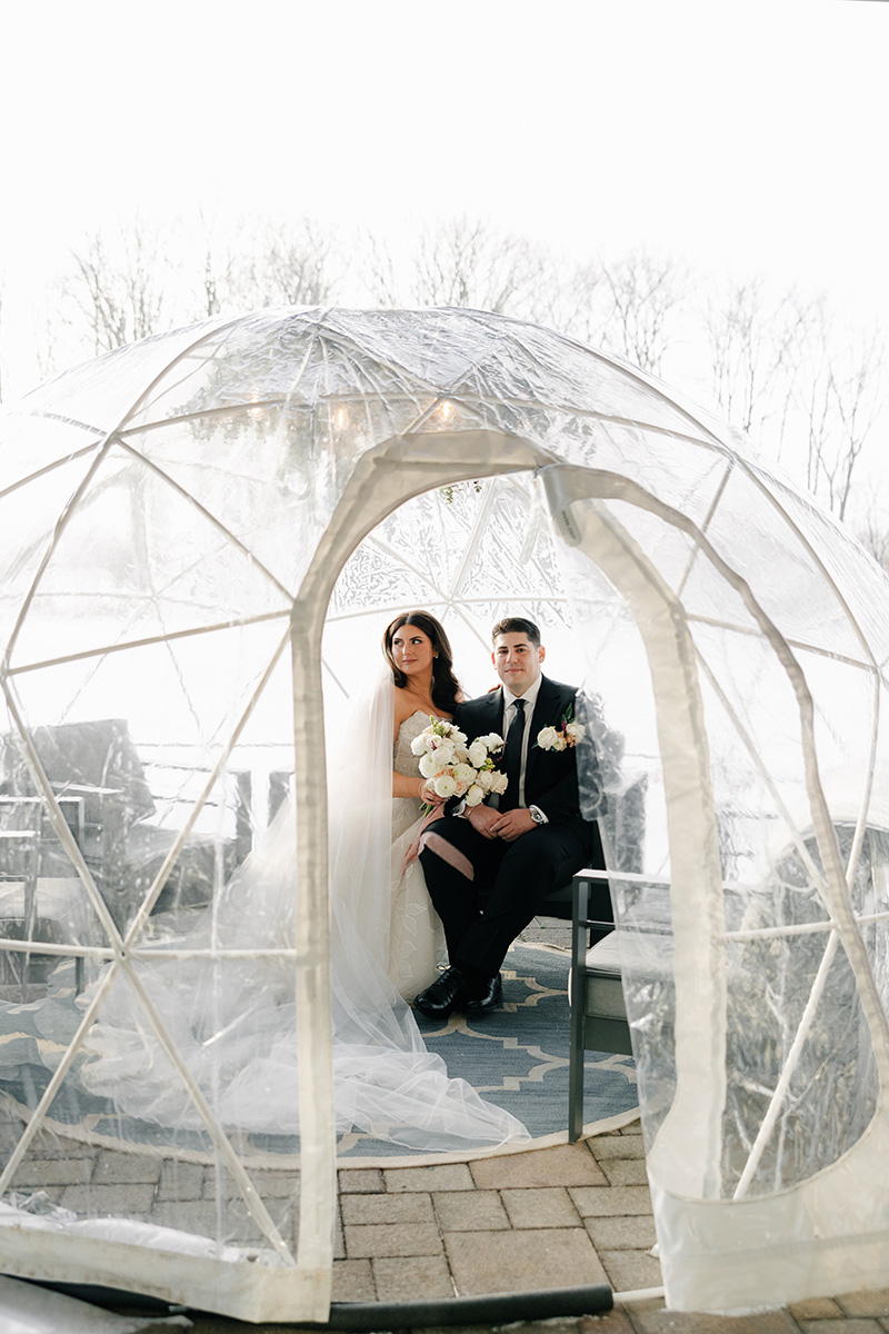Bride and groom sit inside a clear geodesic tent outdoors, winter trees in the background, bouquets in hand.