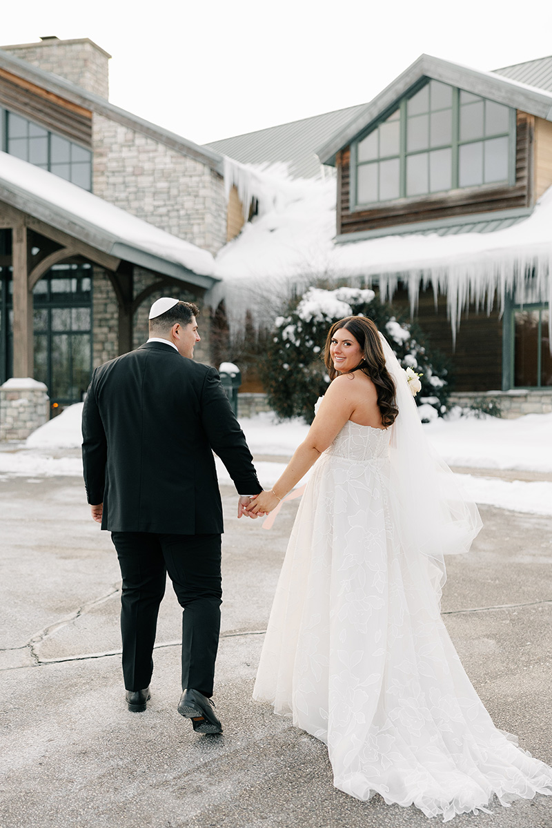 Bride in a white wedding gown and veil holding hands with a groom in a black suit as they walk away from the camera toward a snow-dusted lodge.